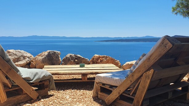 Un pontile di legno con due poltrone da relax e un tavolino, con vista su un mare cristallino e una costa in lontananza.