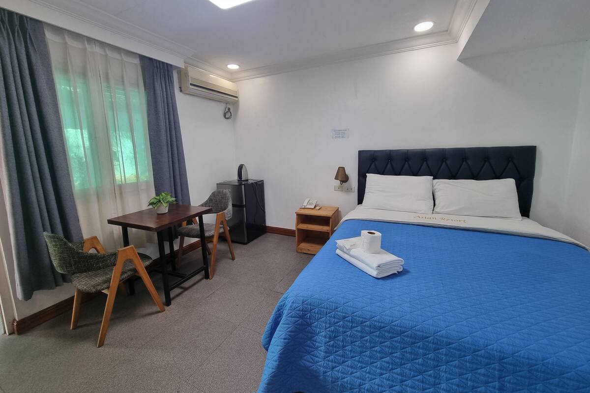 A hotel room with a bed, a desk, a chair, a small table, and a wall-mounted air conditioner.
