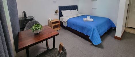 Habitación de hotel con cama, escritorio, silla, mesita con una planta, teléfono en la pared y un cuadro.