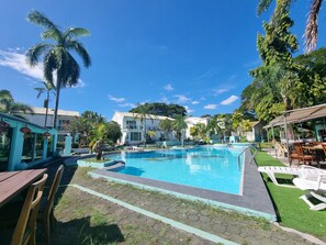 A swimming pool with a tiled edge, surrounded by palm trees and a building with a white facade. -  Asian Greenville Hotel and Resort (Mabalacat City)