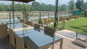 A glass-top table with wicker chairs, a patio umbrella, and a view of a river and mountains. - Upani Cottages (Srinagar)