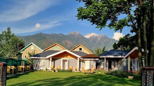 A row of three-story buildings with a mountain backdrop, a well-maintained lawn, and outdoor seating area. - Upani Cottages (Srinagar)