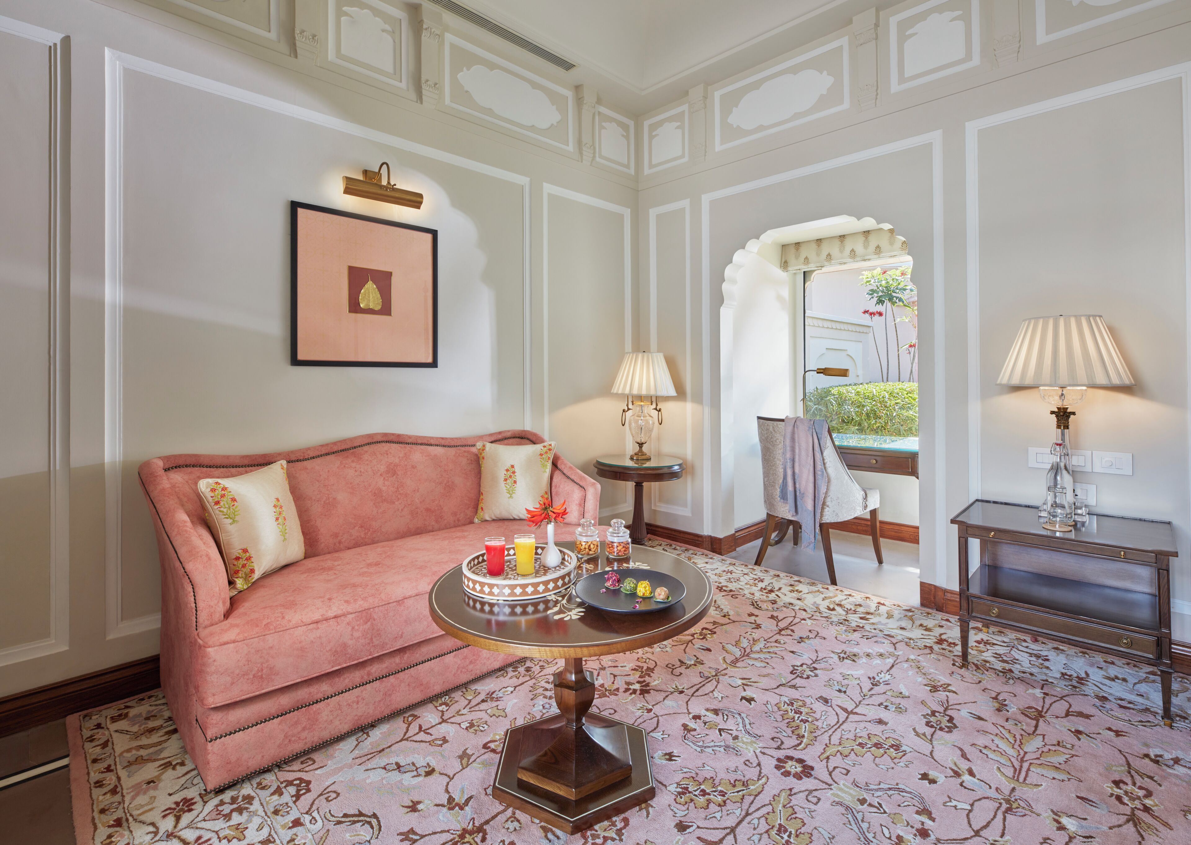 Una habitación con un sofá rosa, una mesa redonda con bebidas, una lámpara y una silla.