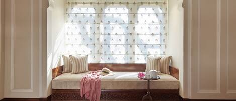 A cozy seating area with a cushioned bench, a small table with a teapot and cup, and a window with patterned curtains.