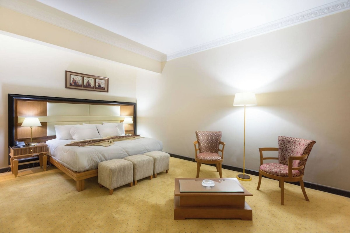 A hotel room with a bed, two armchairs, a floor lamp, a bedside table, and a small table with a lamp.