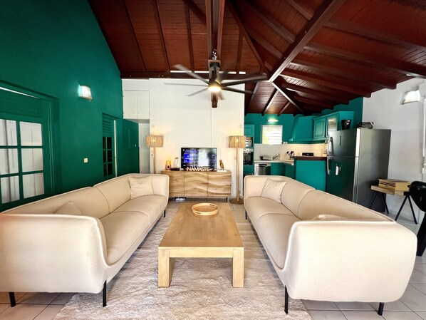 A living room with two sofas, a coffee table, and a TV. There is a kitchen area with teal cabinets and a refrigerator. - Villa Siméon Saint François (Saint-François)