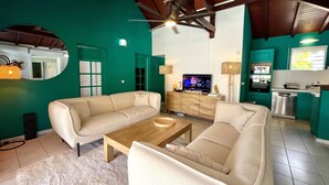 Uma sala de estar com dois sofás bege, uma mesa de centro de madeira, uma TV sobre um móvel de madeira e um ventilador de teto.