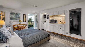 A bedroom with a bed, a small table with chairs, a kitchen area with cabinets and appliances, and a balcony with a view.