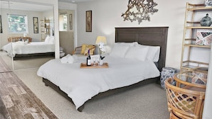 A neatly arranged bedroom with a large bed, a bedside table with a bottle and glasses, a chair, and a decorative wall piece.