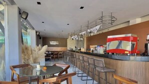 Un restaurante moderno con mesas y sillas de madera, una zona de bar con un equipo rojo y blanco, y un amplio ventanal que proporciona luz natural.