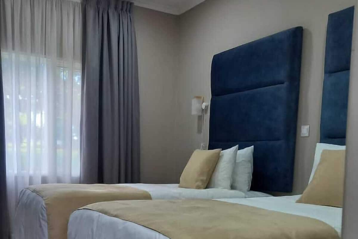Habitación de hotel con dos camas, ropa de cama color crema y blanca, un cabecero azul grande y una ventana con cortinas traslúcidas.