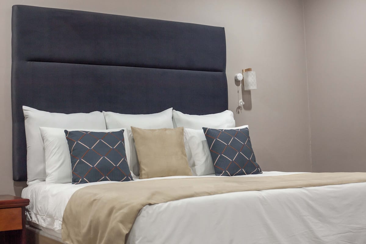 Una cama bien tendida con un cabecero oscuro, almohadas blancas con diseño y una manta color beige.