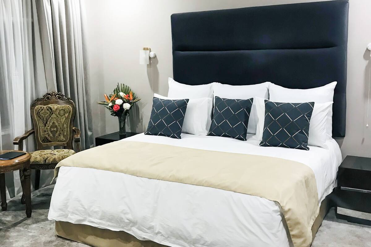 Una cama bien hecha con un cabecero oscuro, ropa de cama blanca y almohadas estampadas. Hay una silla de madera y una mesita de noche con un florero al lado de la cama.