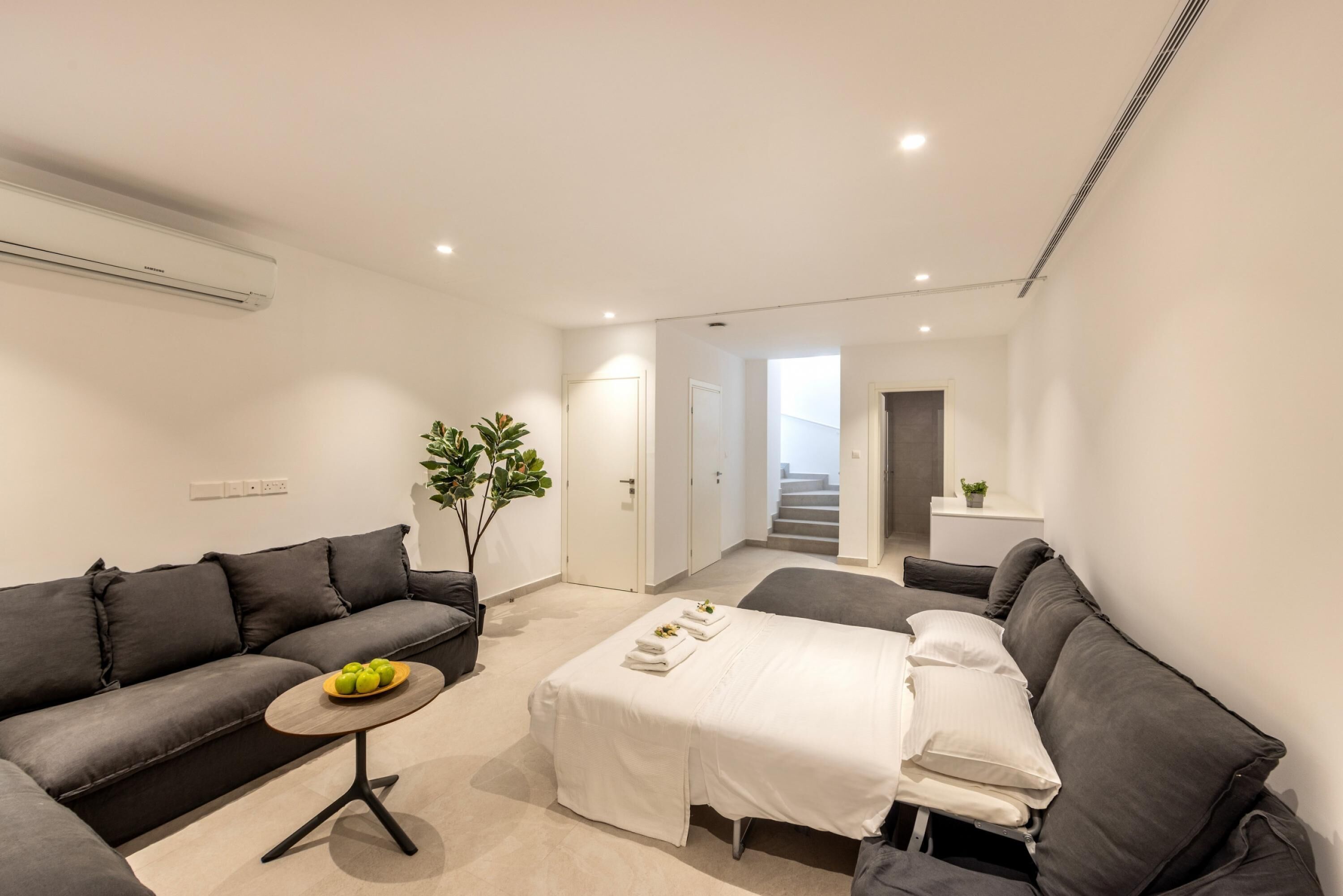 A modern, well-lit room with a grey sofa, a small round table with a bowl of fruit, a bed with white linens, and a staircase leading to an upper level.