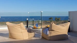 A glass-enclosed balcony with beige cushions, a small round table with drinks, and a clear view of the sea. - Protaras Periyiali Seaside Villa 33 (Paralimni)