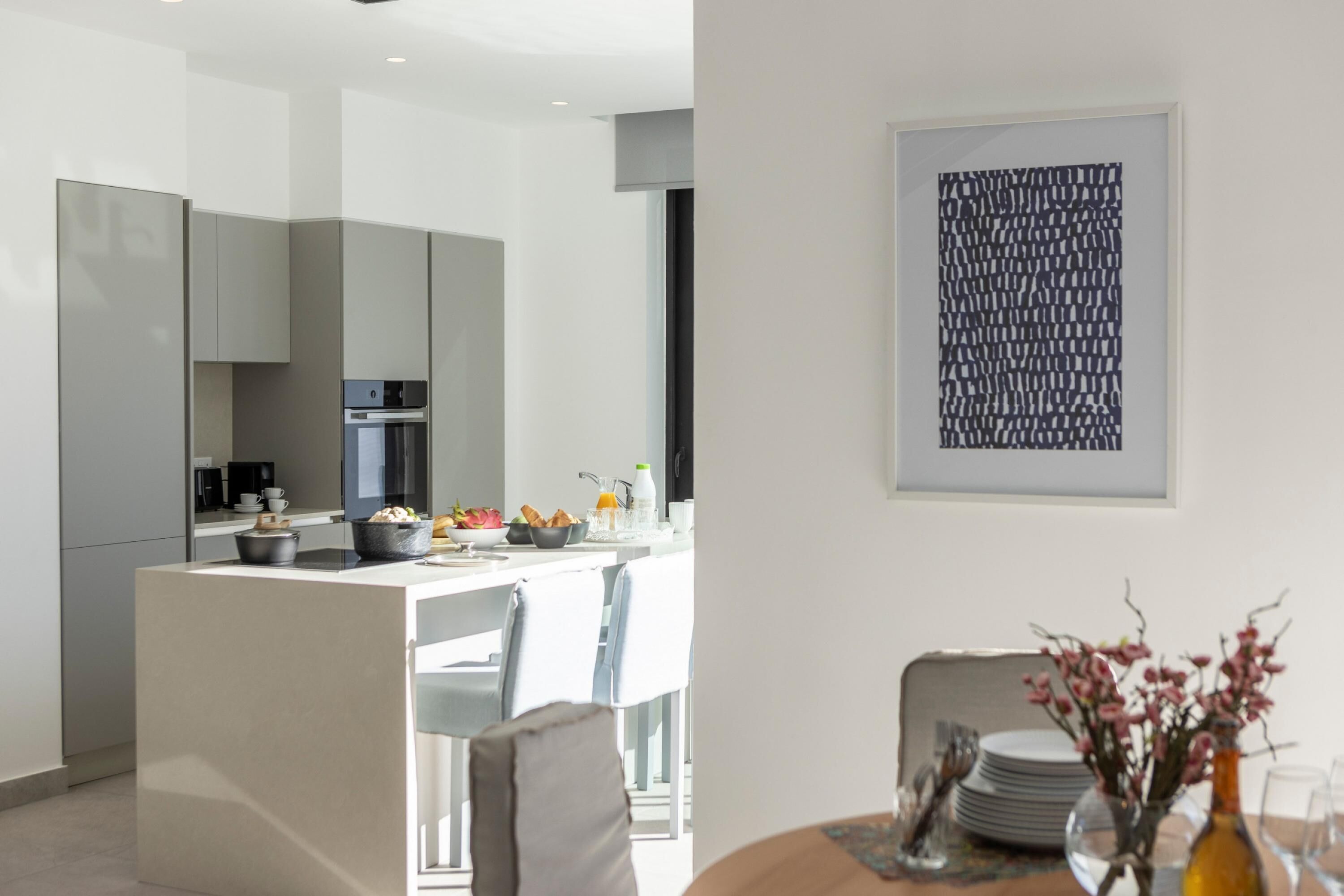 A modern kitchen with white cabinetry, a dining area with a table set for two, and a framed abstract artwork on the wall.