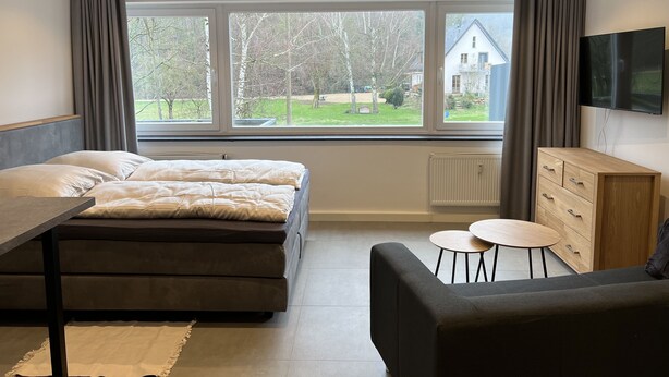 A modern bedroom with a large bed, a TV mounted on the wall, a small table, and a view of the outside. - Apartment 170 (Bad Münstereifel)
