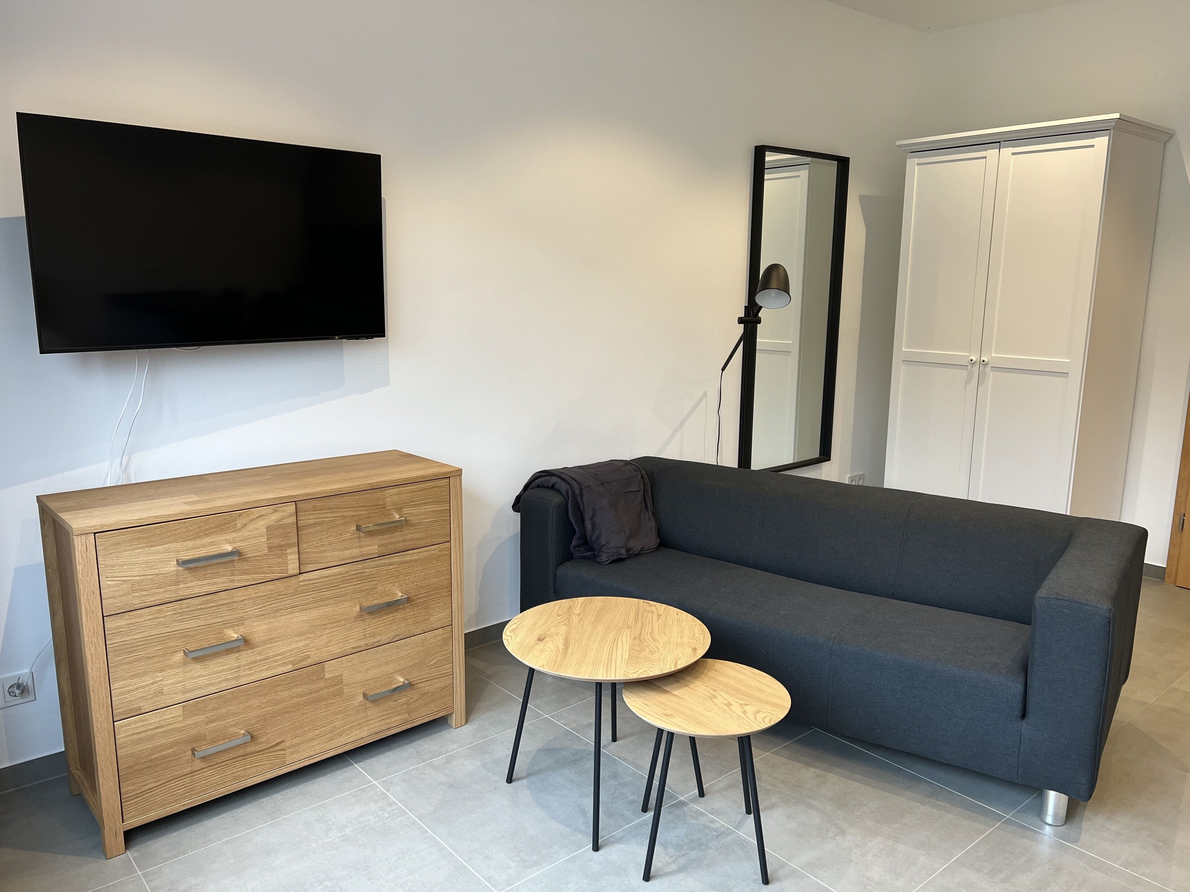 A modern room with a wooden dresser, a grey sofa, two round wooden tables, a flat-screen TV, a mirror, and a wardrobe.