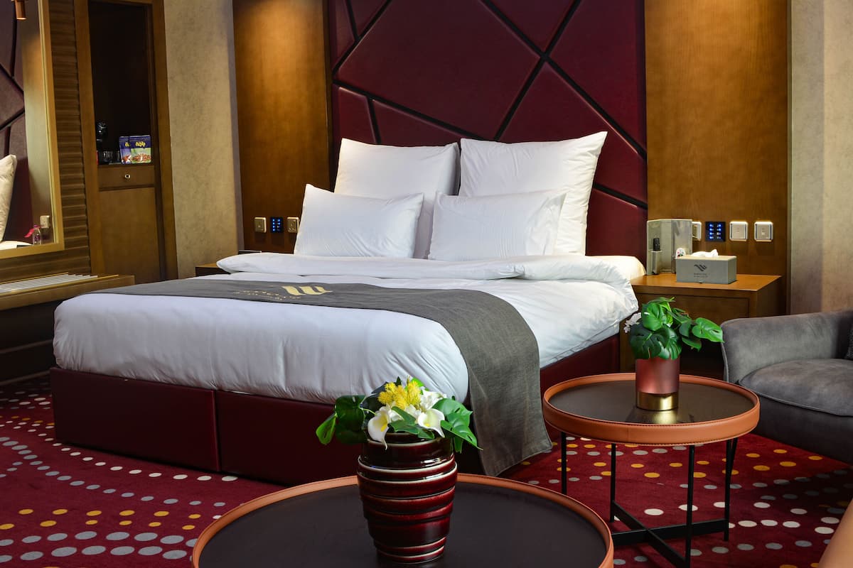Una habitación de hotel moderna con una cama grande, una mesita con un florero, un sillón gris y una alfombra roja con estampado de lunares.