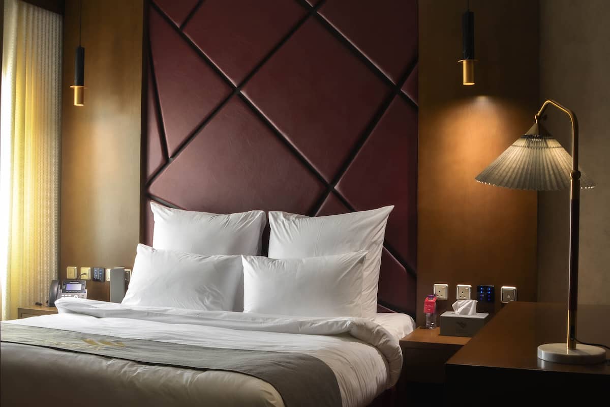 Una cama bien hecha con ropa blanca, una mesita de noche con una lámpara y una pared con un diseño geométrico.