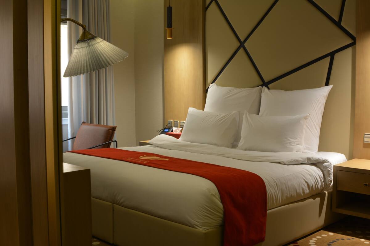 Una habitación de hotel con cama, mesita de noche, lámpara, silla y ventana con cortinas.