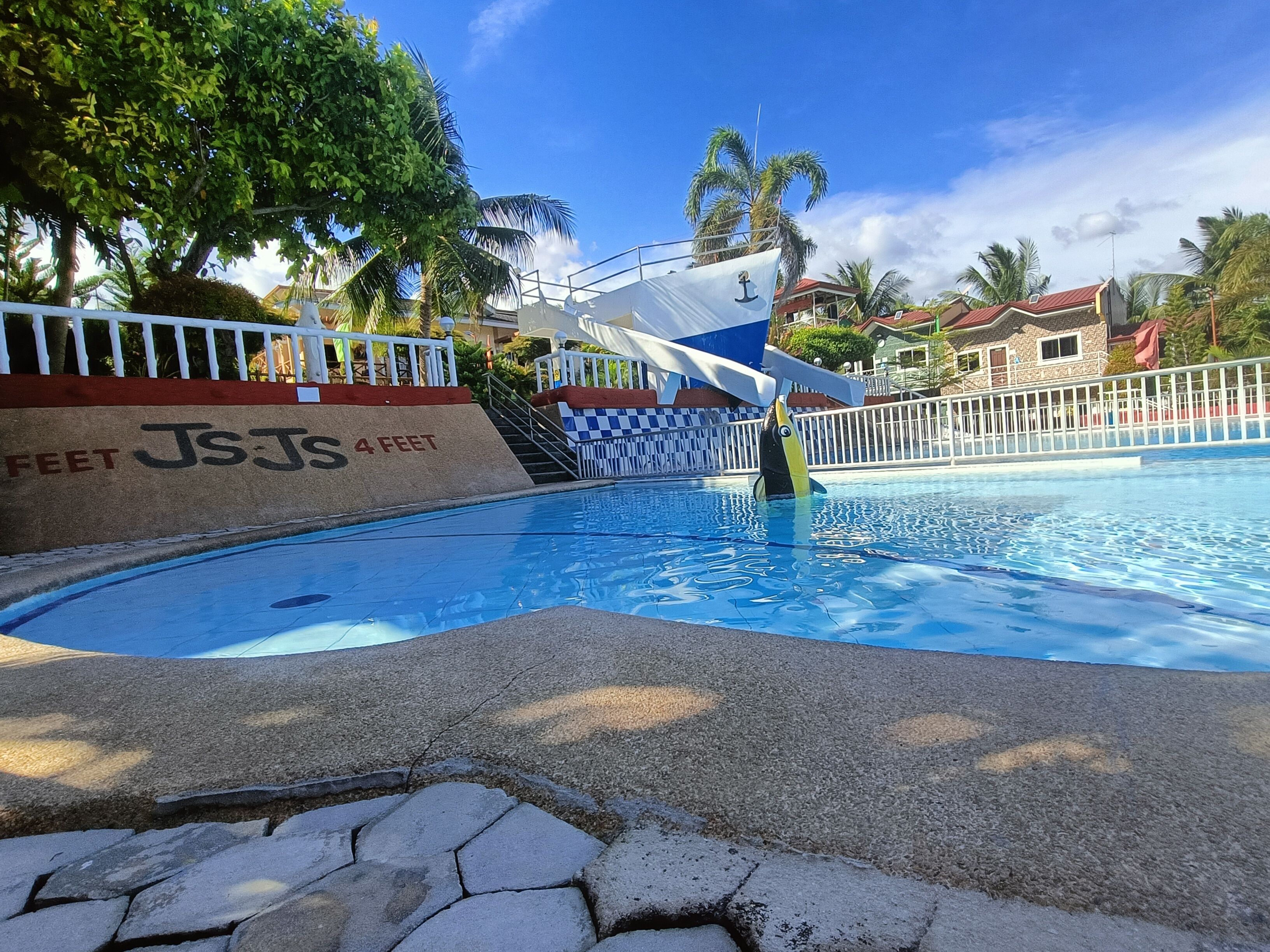 Una piscina con una torre de salvavidas, palmeras y un edificio de techo rojo al fondo.