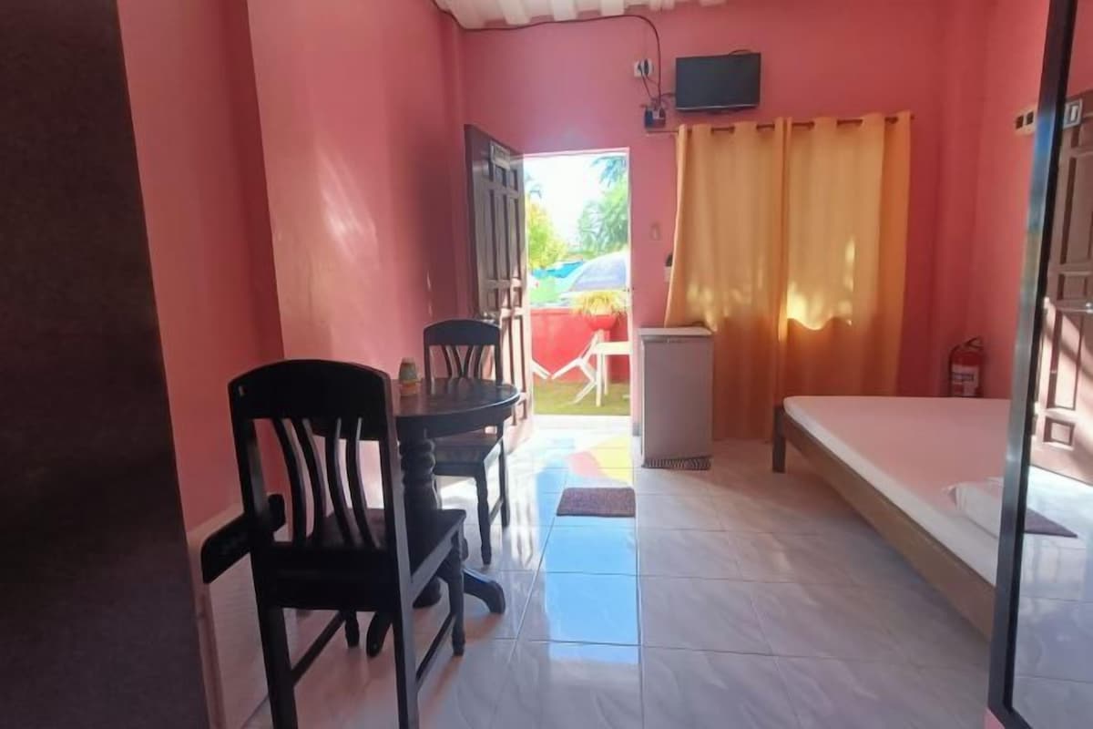 A room with a bed, a table with chairs, a small refrigerator, and a window with curtains.