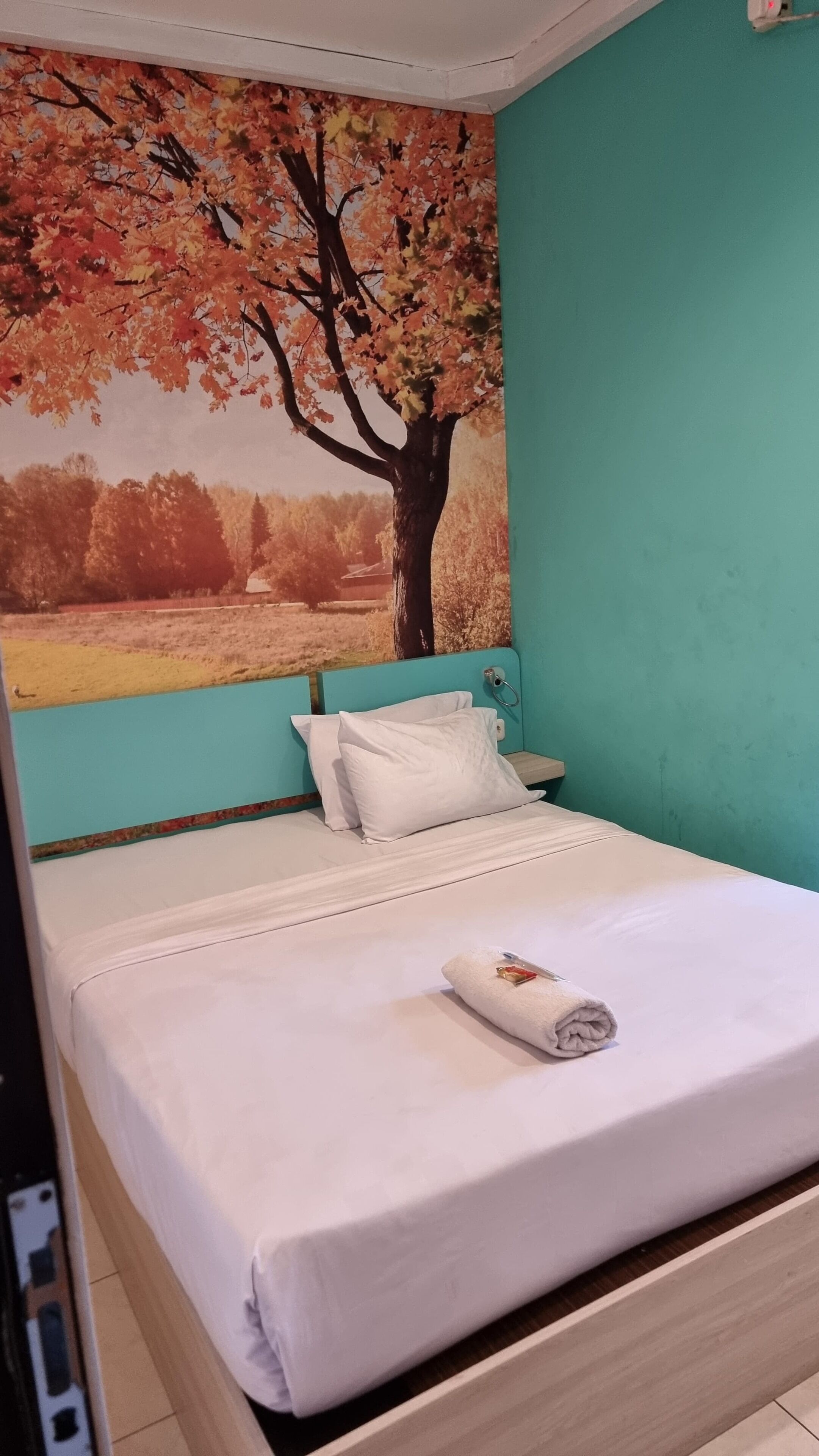 Un dormitorio con cama, ropa de cama blanca, una pared verde azulado y un mural grande de un árbol otoñal.