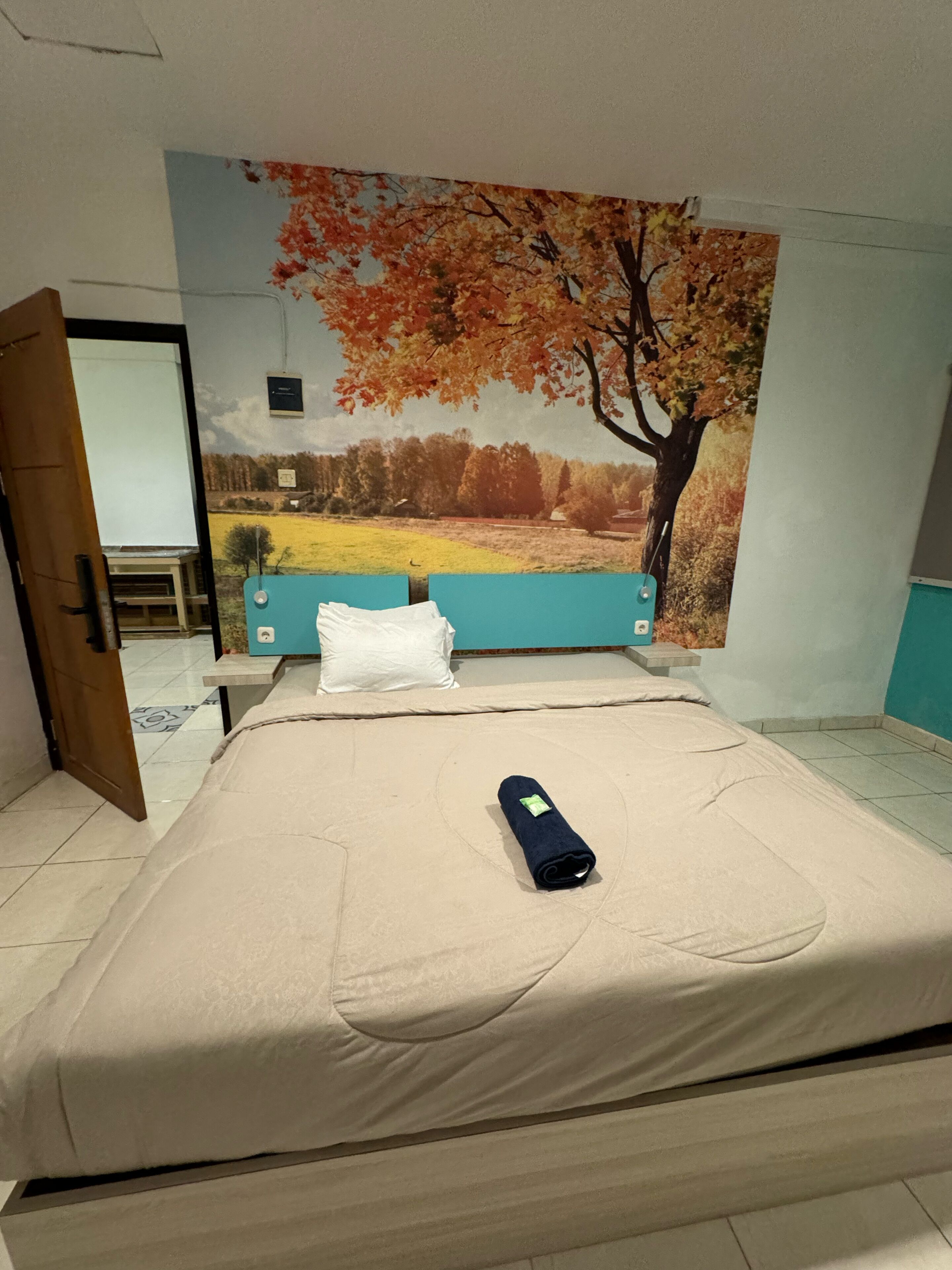 A neatly made bed with a beige mattress, a rolled-up dark blue towel, and a white pillow. A large landscape painting of autumn trees and a field is on the wall behind the bed.