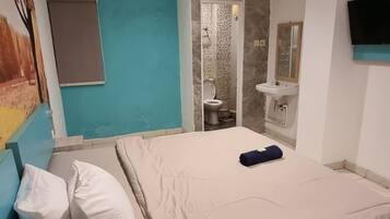 Un dormitorio con cama, almohadas, una mesita de noche, un espejo, un televisor y un baño con inodoro y lavabo.