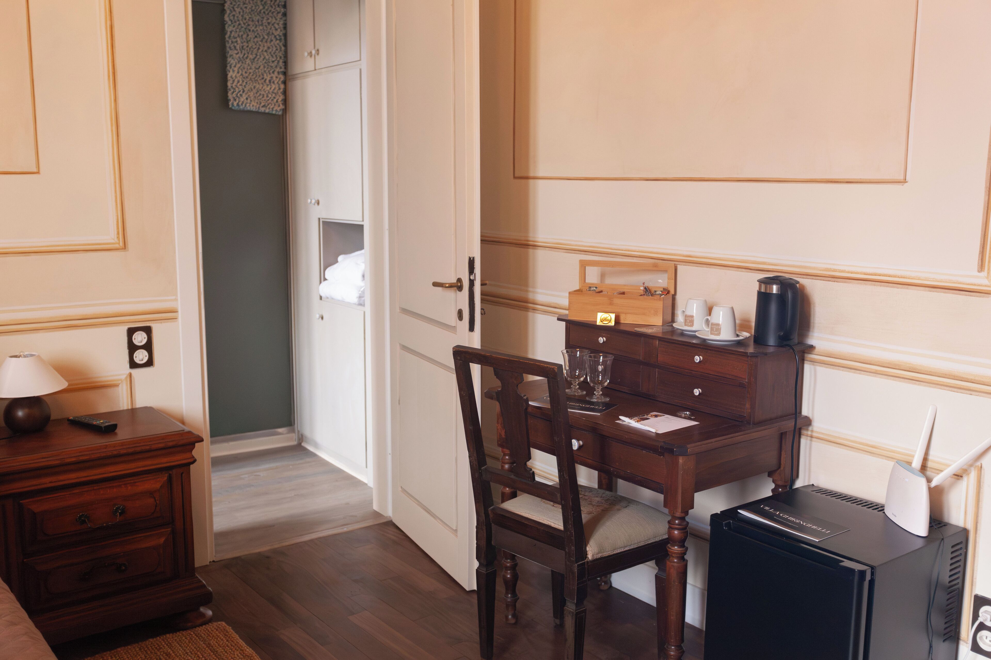Habitación de hotel con escritorio de madera, una silla, una lámpara, una cafetera y un pequeño dispositivo negro en el suelo.