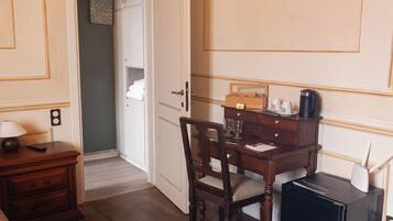 Habitación de hotel con escritorio de madera, una silla, una lámpara, una cafetera y un pequeño dispositivo negro en el suelo.