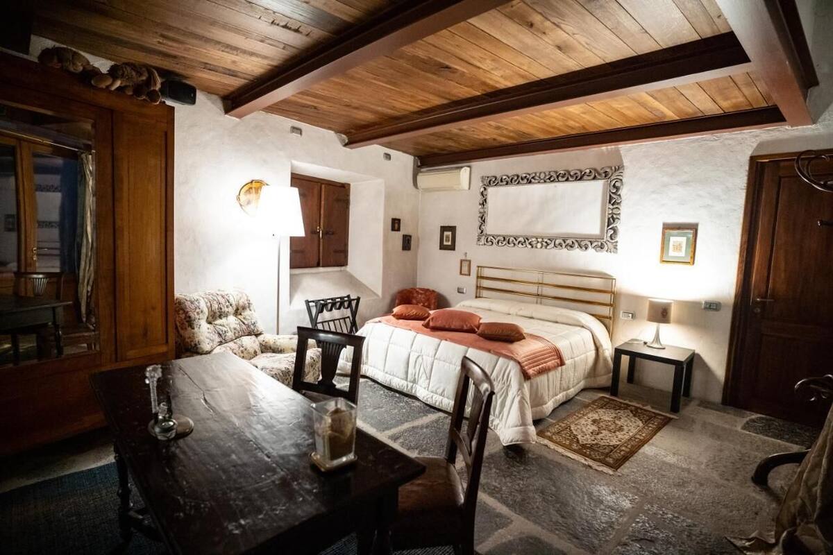 A cozy bedroom with a wooden ceiling, a bed with a white headboard, a small table with chairs, and a rug on the floor.