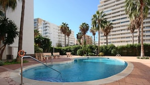 Une piscine équipée d’une bouée de sauvetage, entourée de palmiers et de bancs, avec des bâtiments en arrière-plan.