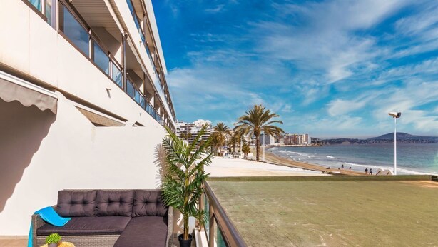 Un balcon avec un canapé, une petite table et un pot de fleurs. Le balcon donne sur une plage bordée de palmiers et offre une vue sur la mer.