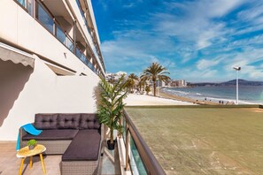Ein Balkon mit einer Couch, einem kleinen Tisch und einer Topfpflanze. Der Balkon bietet Blick auf einen Strand mit Palmen und Meer.