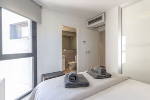 Habitación de hotel moderna con un ventanal grande, una cama con dos toallas enrolladas, una mesita de noche pequeña y un baño visible con inodoro y ducha.