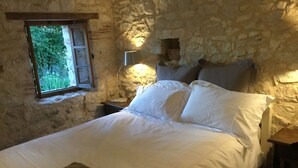 A bedroom with a stone wall, a bed with white linens, a bedside lamp, and a window with a view of greenery.