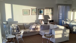 A living room with a beige sofa, a small round table, and a lamp. There are framed pictures on the wall and a window with curtains. - Ile aux Moines (Arzon)
