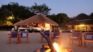 Un salón frente a la playa con estructuras de techo de paja, sillas de madera y un pozo central para fuego.