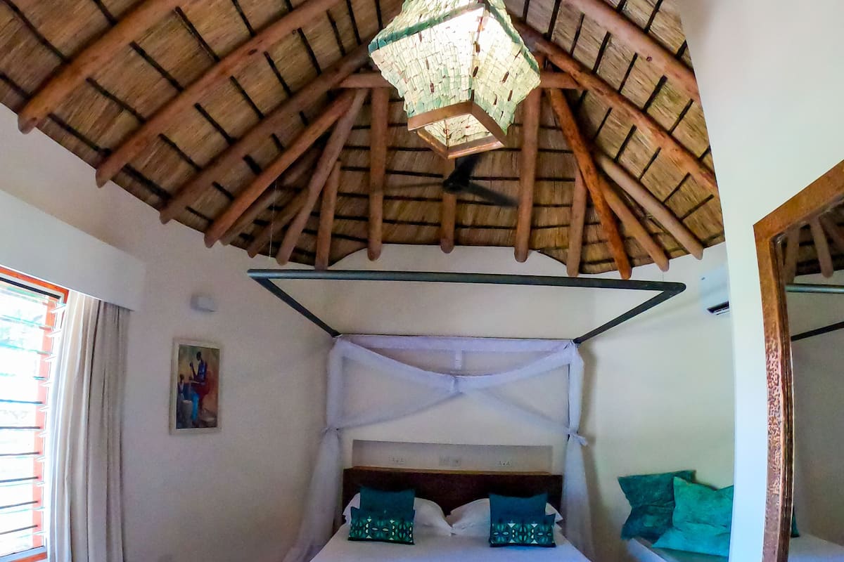 Een kamer met een hoog houten plafond, een bed met witte beddengoed en turquoise kussens, een hanglamp en een schilderij aan de muur.