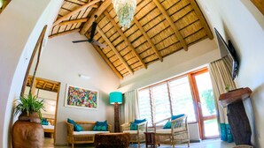 A spacious living room with a high ceiling, wooden beams, a chandelier, a large window, wicker furniture, and a flat-screen TV.