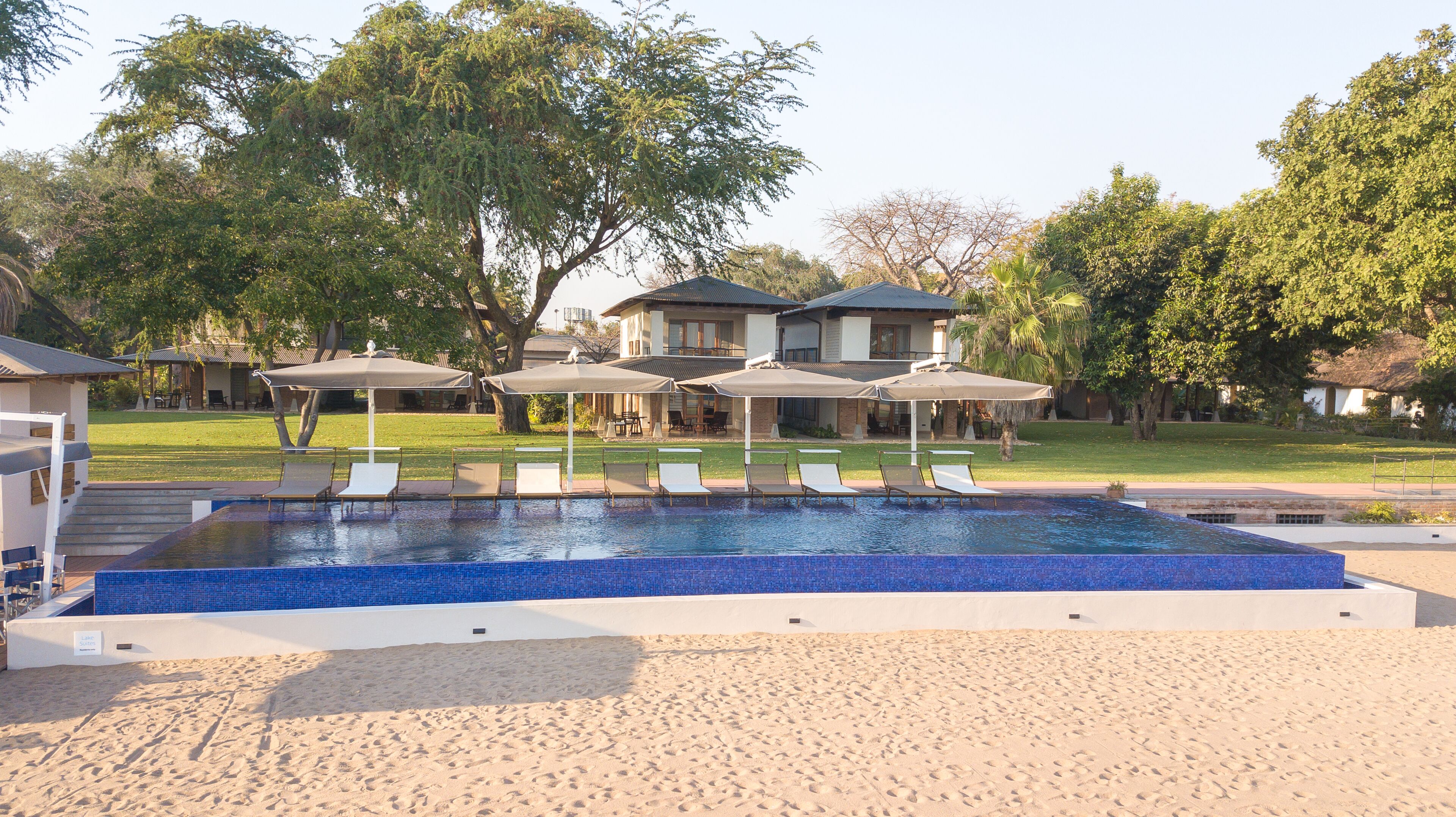 Una piscina con sillones de playa y sombrillas, rodeada de arena y árboles.