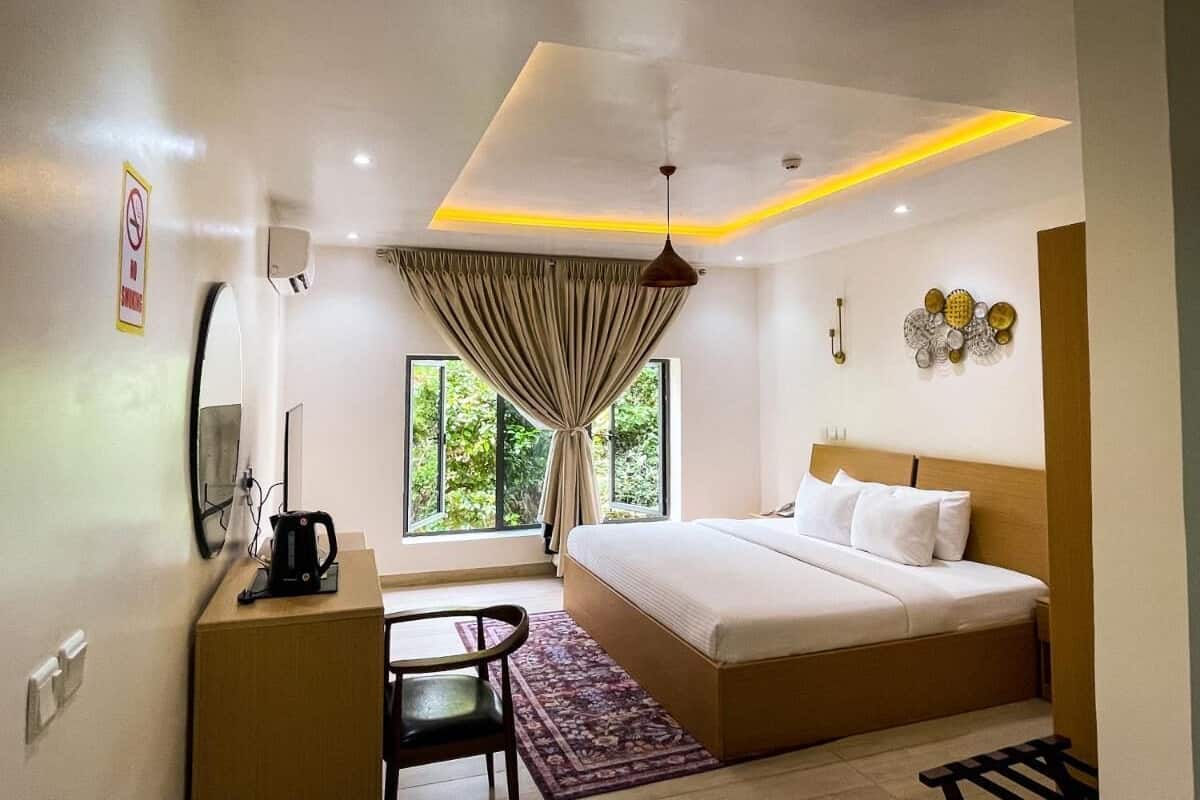 A modern hotel room with a large bed, a desk with a kettle, a chair, and a view of greenery through the window.