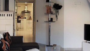 A modern living room with a white sofa, a white ottoman, a glass coffee table, and a black sofa. There is a kitchen in the background with a stove and cabinets. - Corse LES Pieds Dans L'eau (Borgo)