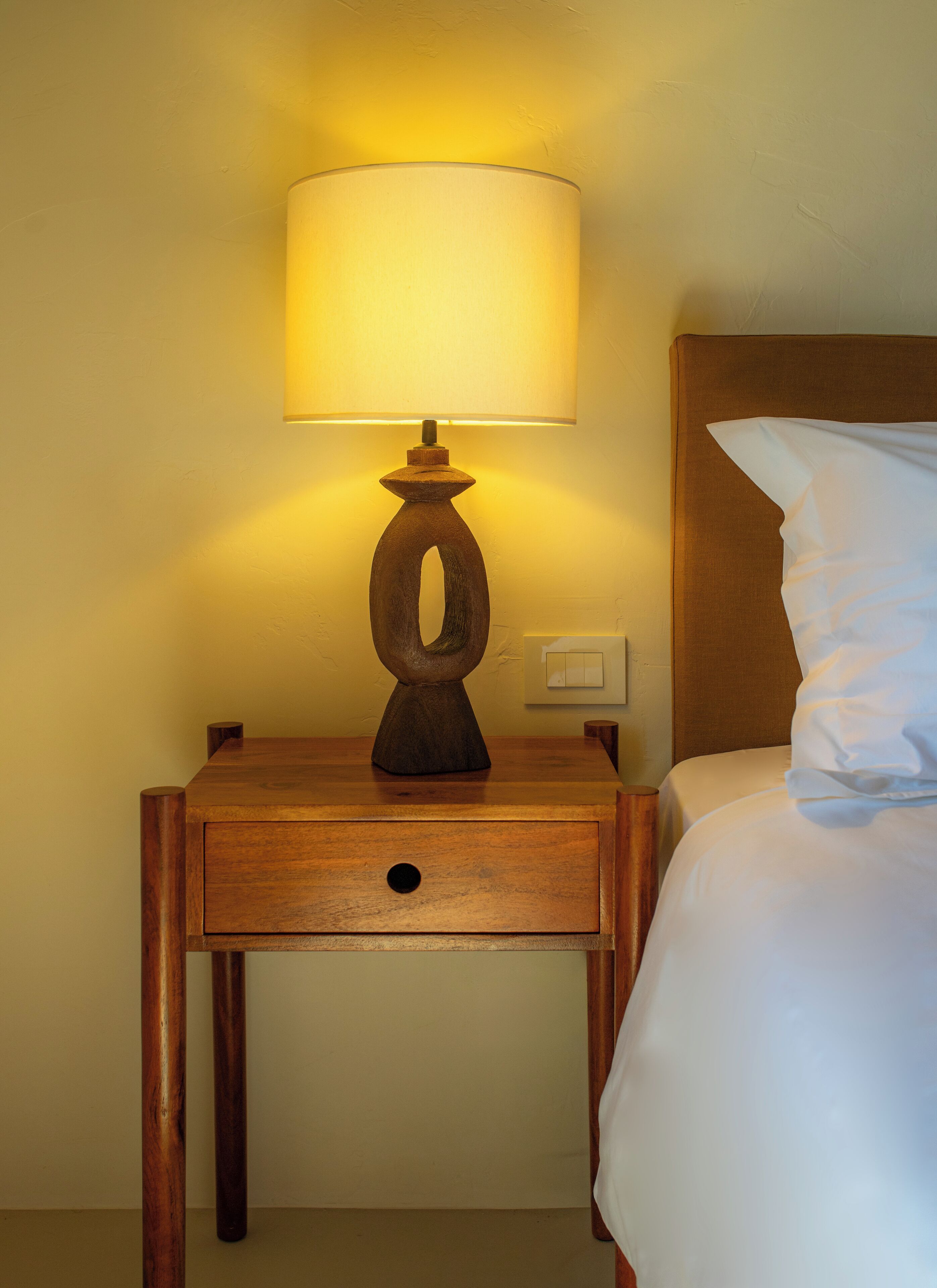 A wooden bedside table with a lamp, a drawer, and a bed with white bedding.