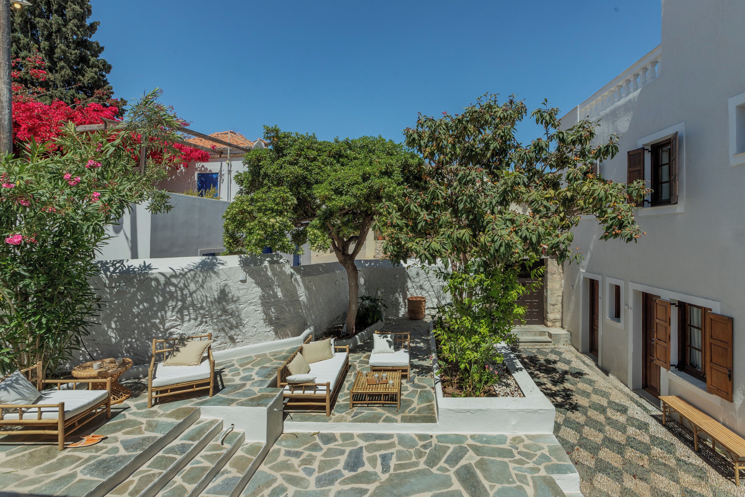 A courtyard with stone flooring, outdoor seating, and a view of a white building with wooden shutters.