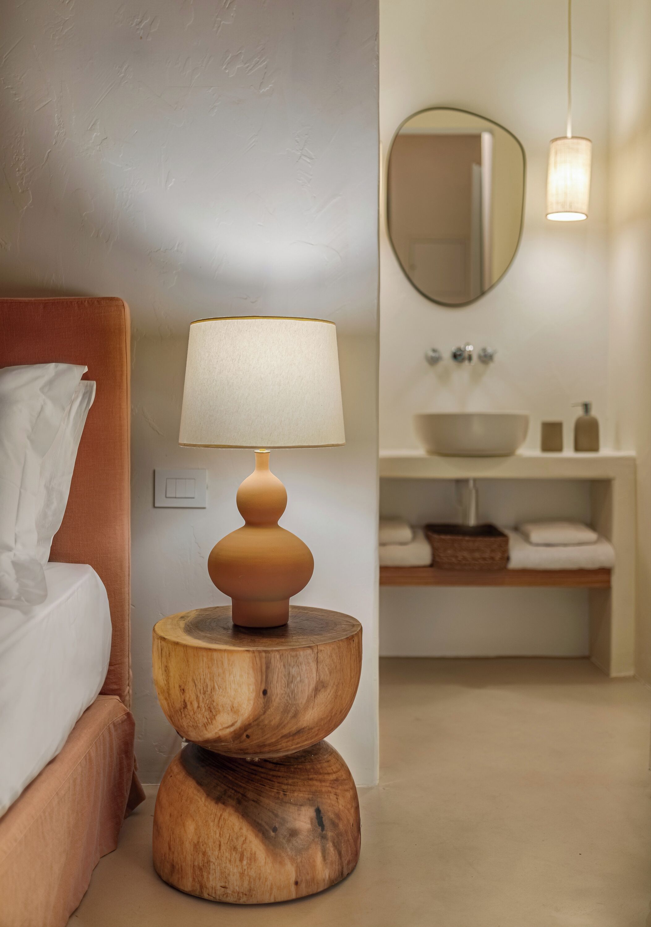 Une chambre à coucher avec une table de chevet en bois, une lampe, un lit et une salle de bain comprenant un lavabo et un miroir.