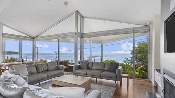 A spacious living room with a large glass wall offering a view of the ocean. There are two leather sofas, a coffee table, and a flat-screen TV mounted on the wall. - A Beachside Getaway 12 by HolidayCo. (KILLCARE)