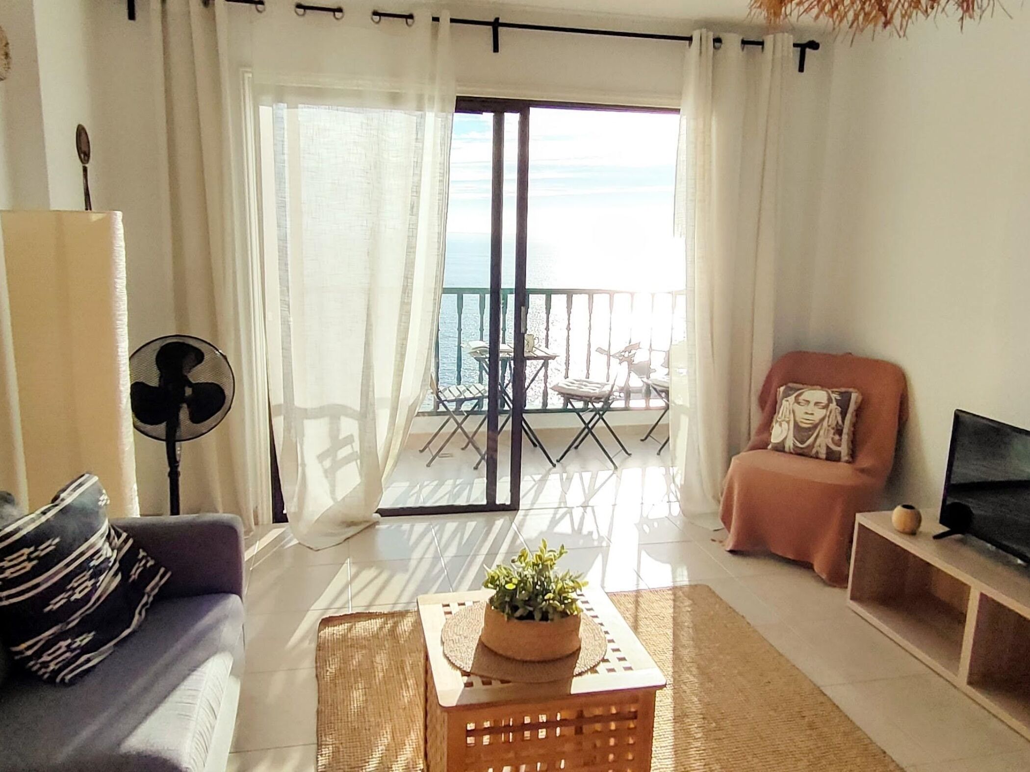 A living room with a sofa, a coffee table with a plant, a TV stand, and a balcony with outdoor furniture.